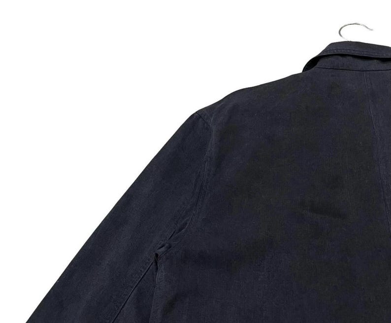 May include: A navy blue jacket with a collar and button closure, hanging on a metal hanger. The jacket's fabric appears to be a sturdy material, and the garment is viewed from the shoulder and upper arm.
