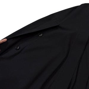 May include: Black coat with two buttons and a notched lapel. The coat is made of a smooth, dark fabric. The image shows a close-up of the coat's front, with a hand partially visible.