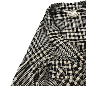 May include: Close-up of a black and white patterned jacket. The fabric has a classic houndstooth and plaid design. The jacket has a frayed edge and a label that reads "LIMI".