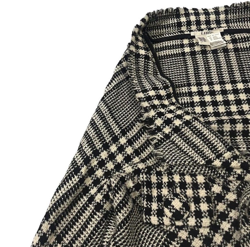 May include: Close-up of a black and white patterned jacket. The fabric has a classic houndstooth and plaid design. The jacket has a frayed edge and a label that reads "LIMI".