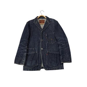 Può includere: Giacca di jeans blu scuro in stile blazer. Presenta un colletto a revers, due tasche frontali e una chiusura con bottoni. La giacca è appesa a una gruccia di legno su sfondo bianco.