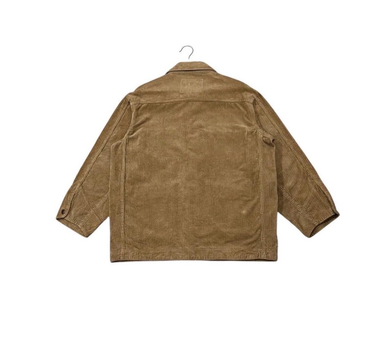 K&ouml;nnte beinhalten: Eine hellbraune Cordjacke, von hinten gesehen, h&auml;ngt an einem Metallhaken. Die Jacke hat einen Knopfverschluss, zwei Brusttaschen und lange &Auml;rmel. Der Stoff hat eine vertikale Rippenstruktur.