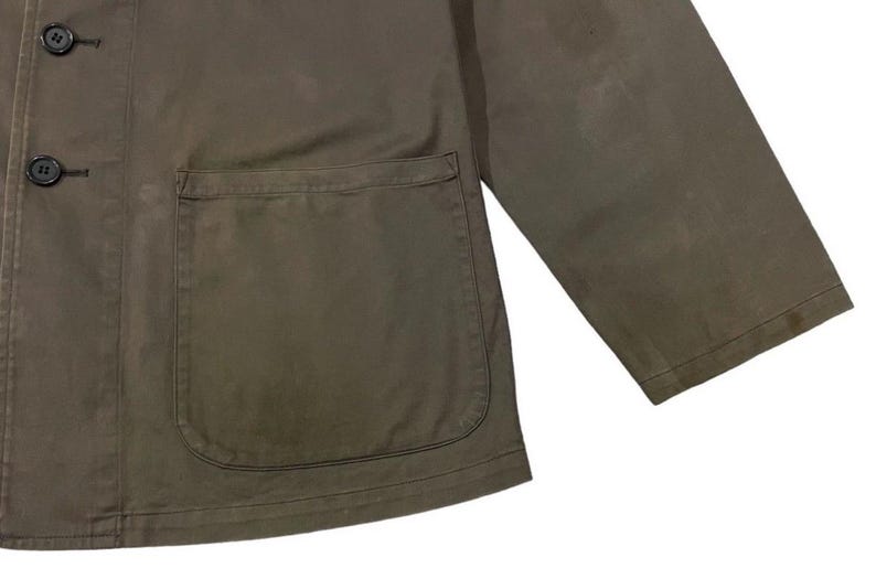 May include: Olive green jacket with a large front pocket and black buttons. The jacket has a classic design with a simple, functional aesthetic. The fabric appears to be a durable material, suitable for various weather conditions.