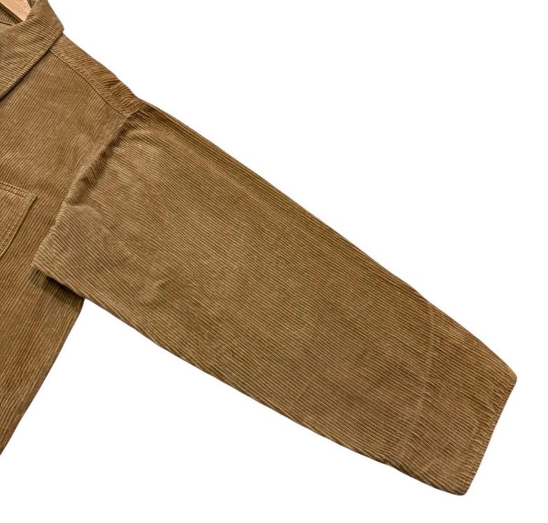K&ouml;nnte beinhalten: Nahaufnahme eines tan-farbenen Cordjacken&auml;rmels. Der Stoff hat eine gerippte Textur und einen warmen, erdigen Ton. Die Jacke scheint neu zu sein, mit einem strukturierten Design und einer klassischen &Auml;sthetik. Der &Auml;rmel ist abgewinkelt und zeigt die Richtung des Stoffes.