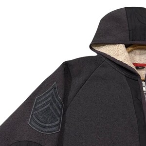 May include: A dark grey hooded sweatshirt with a black panel down the front and a patch on the left sleeve. The hood is lined with a cream-coloured, fuzzy material. The sweatshirt has a zip closure.