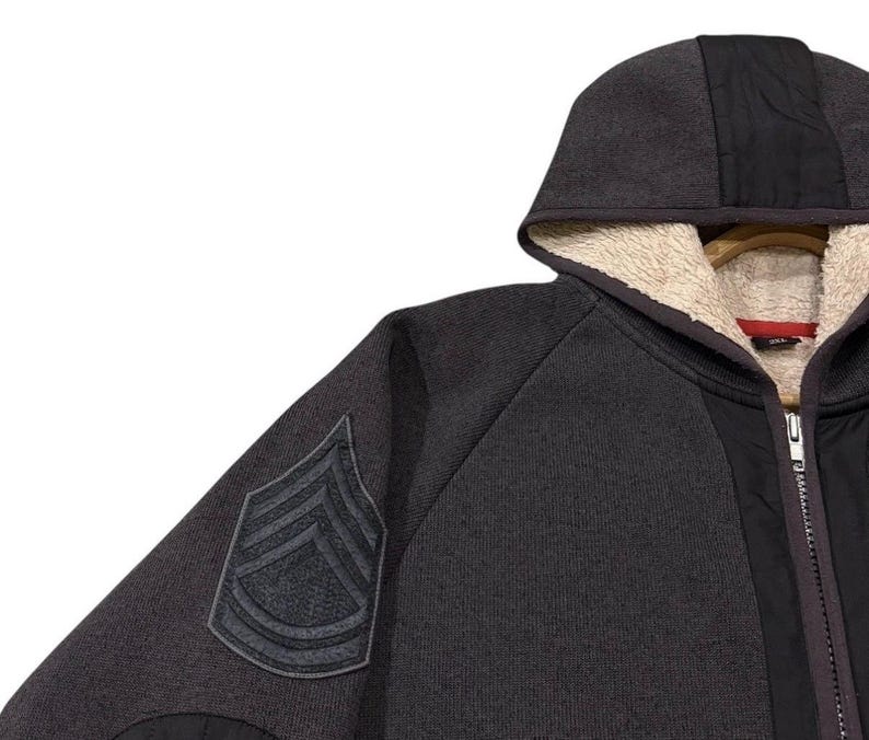 May include: A dark grey hooded sweatshirt with a black panel down the front and a patch on the left sleeve. The hood is lined with a cream-coloured, fuzzy material. The sweatshirt has a zip closure.