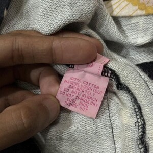 May include: A pink clothing label with black text, held by a hand, against a grey and black striped fabric. The label reads "100% COTTON MACHINE WASH COLD SEPARATELY DO NOT BLEACH DO NOT TUMBLE DRY COOL IRON INSIDE OUT."