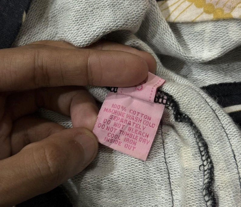 May include: A pink clothing label with black text, held by a hand, against a grey and black striped fabric. The label reads "100% COTTON MACHINE WASH COLD SEPARATELY DO NOT BLEACH DO NOT TUMBLE DRY COOL IRON INSIDE OUT."