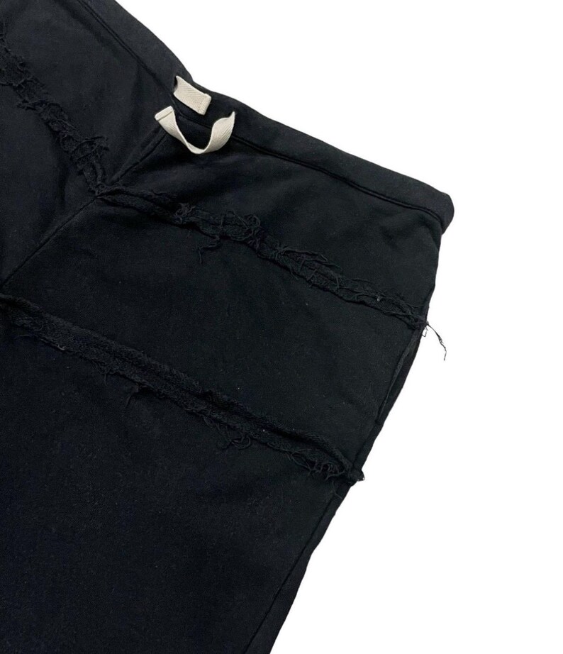 Puede incluir: Primer plano de un pantal&oacute;n de ch&aacute;ndal negro con detalles desgastados. El pantal&oacute;n tiene una cintura con cord&oacute;n y varias costuras horizontales desgastadas en la parte delantera. La tela parece ser un material suave y oscuro.