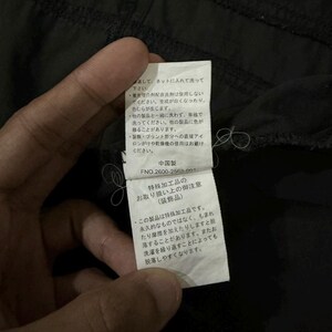 Puede incluir: Primer plano de una etiqueta de prenda negra sujeta por una mano. La etiqueta tiene texto japon&eacute;s y un c&oacute;digo de producto. La etiqueta proporciona instrucciones de cuidado de la prenda, incluyendo consejos de lavado y planchado.