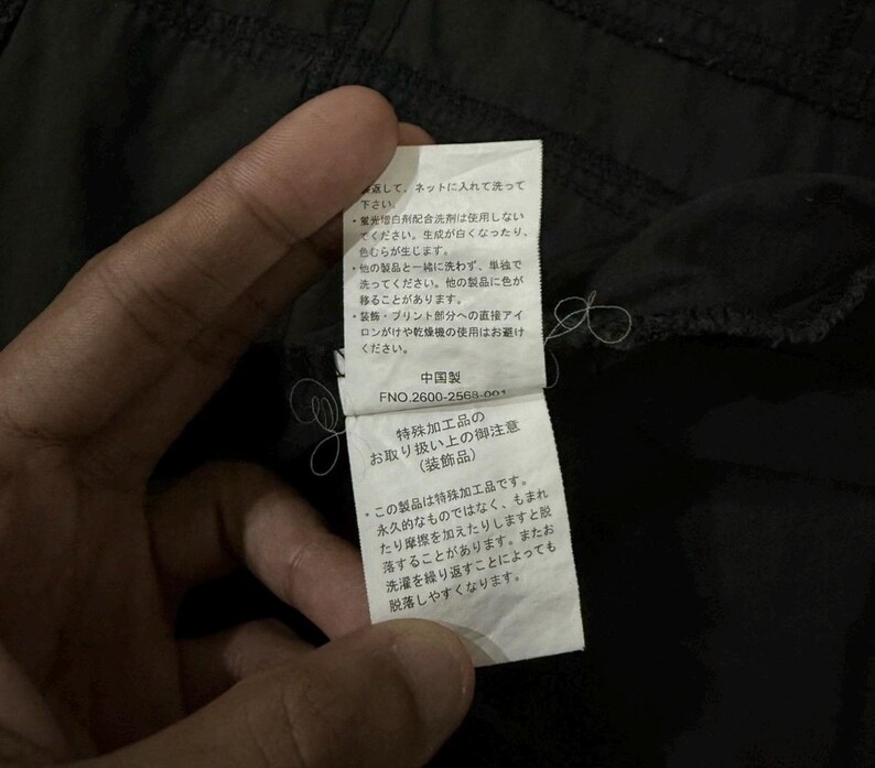 Puede incluir: Primer plano de una etiqueta de prenda negra sujeta por una mano. La etiqueta tiene texto japon&eacute;s y un c&oacute;digo de producto. La etiqueta proporciona instrucciones de cuidado de la prenda, incluyendo consejos de lavado y planchado.