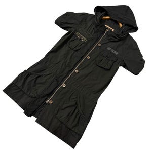 Puede incluir: Chaqueta negra con capucha, mangas cortas, m&uacute;ltiples bolsillos y cierre de cremallera. La chaqueta presenta tachuelas decorativas y el texto "FOX DUTCH". Una prenda de abrigo elegante y funcional.