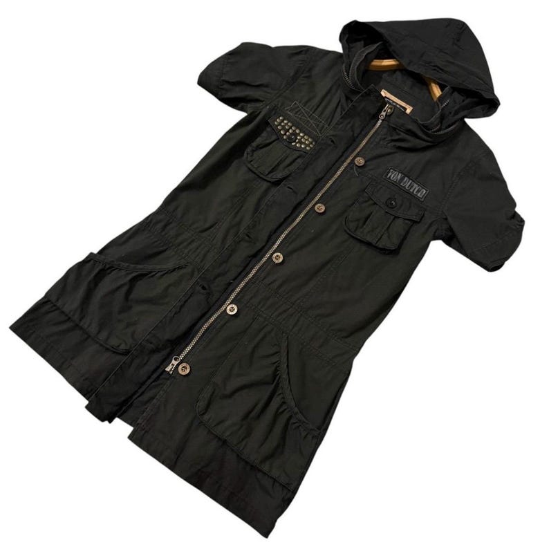 Puede incluir: Chaqueta negra con capucha, mangas cortas, m&uacute;ltiples bolsillos y cierre de cremallera. La chaqueta presenta tachuelas decorativas y el texto "FOX DUTCH". Una prenda de abrigo elegante y funcional.