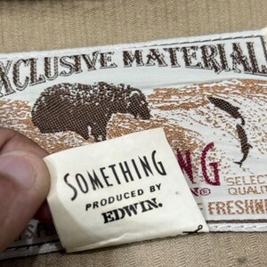 K&ouml;nnte beinhalten: Nahaufnahme eines Stoffetiketts mit den Worten "EXCLUSIVE MATERIAL" und einer B&auml;renillustration. Ein kleineres Etikett lautet "SOMETHING PRODUCED BY EDWIN."