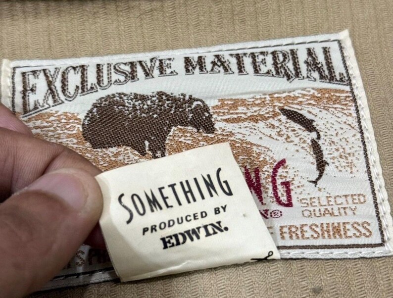 K&ouml;nnte beinhalten: Nahaufnahme eines Stoffetiketts mit den Worten "EXCLUSIVE MATERIAL" und einer B&auml;renillustration. Ein kleineres Etikett lautet "SOMETHING PRODUCED BY EDWIN."
