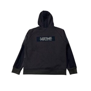 May include: Dark grey hoodie with a hood and long sleeves. The back of the hoodie features a rectangular patch with the text "DE OPPRESSO LIBER - GREEN BERET - U.S. ARMY SPECIAL FORCE". The sleeves have a darker grey panel.