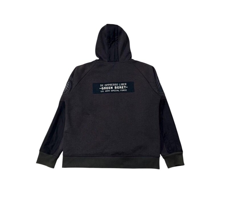May include: Dark grey hoodie with a hood and long sleeves. The back of the hoodie features a rectangular patch with the text "DE OPPRESSO LIBER - GREEN BERET - U.S. ARMY SPECIAL FORCE". The sleeves have a darker grey panel.