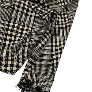 May include: Close-up of a black and white plaid fabric. The fabric has a classic houndstooth pattern with a frayed edge. The material appears to be woven, with a textured surface. The color scheme is monochromatic, with black and white squares.
