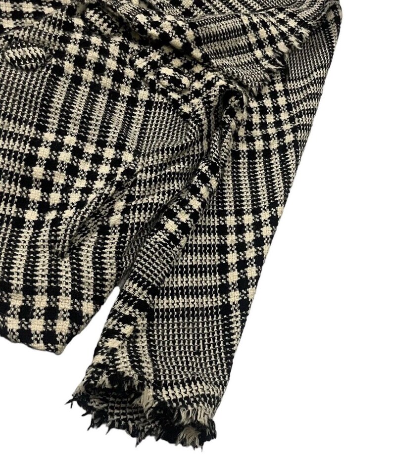 May include: Close-up of a black and white plaid fabric. The fabric has a classic houndstooth pattern with a frayed edge. The material appears to be woven, with a textured surface. The color scheme is monochromatic, with black and white squares.