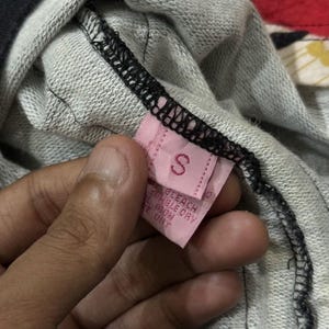May include: A close-up of a pink clothing label with the letter "S". The label is attached to a grey and black garment. The label also has care instructions printed on it.