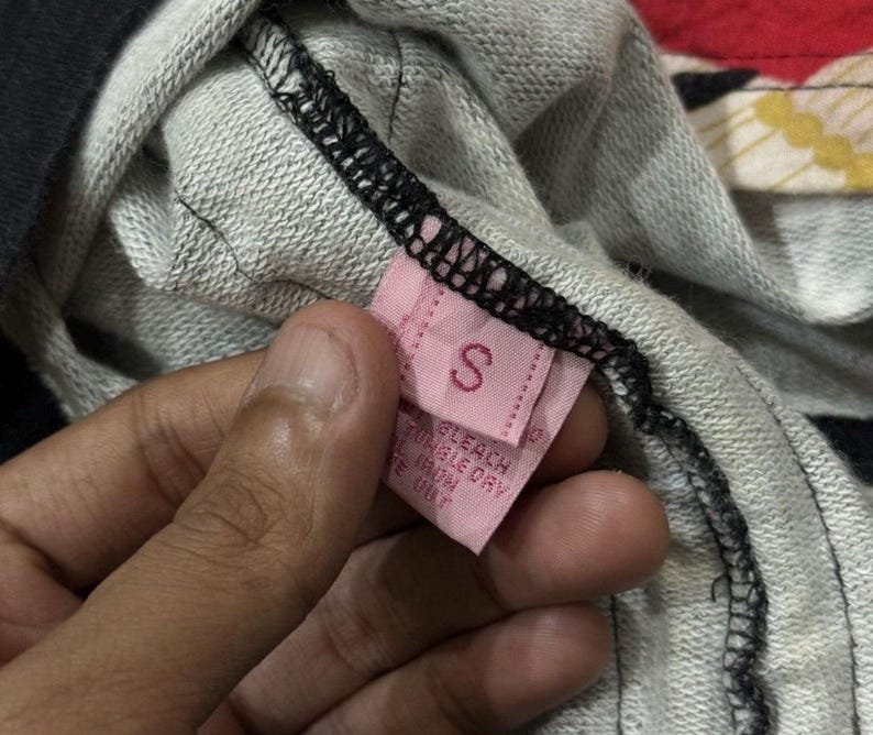May include: A close-up of a pink clothing label with the letter "S". The label is attached to a grey and black garment. The label also has care instructions printed on it.