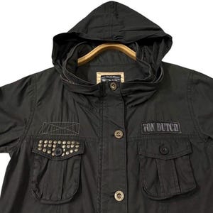 Puede incluir: Chaqueta verde oliva oscuro con capucha y mangas cortas. La chaqueta presenta dos bolsillos delanteros con tachuelas met&aacute;licas, un parche con el texto "VON DUTCH" y cierre de cremallera. La chaqueta est&aacute; colgada en una percha de madera.