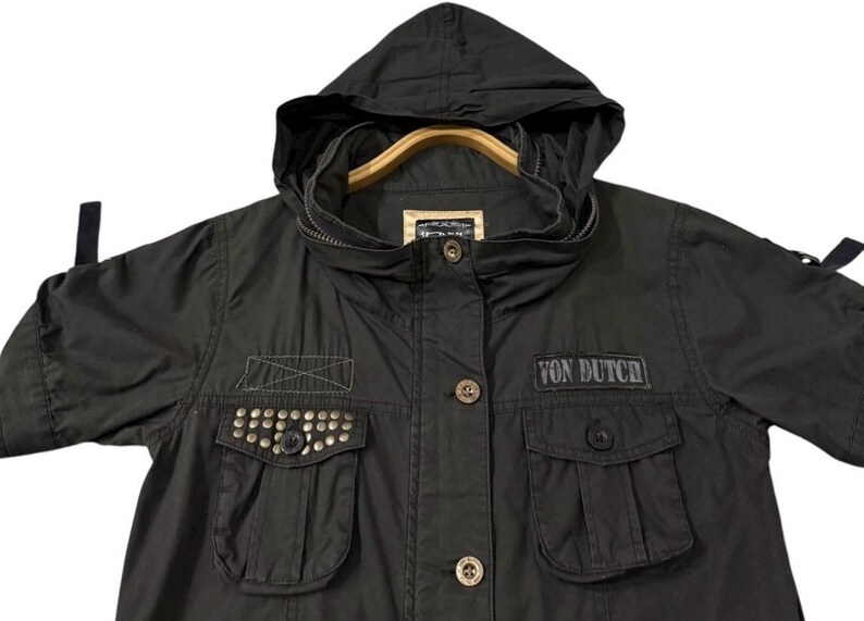 Puede incluir: Chaqueta verde oliva oscuro con capucha y mangas cortas. La chaqueta presenta dos bolsillos delanteros con tachuelas met&aacute;licas, un parche con el texto "VON DUTCH" y cierre de cremallera. La chaqueta est&aacute; colgada en una percha de madera.