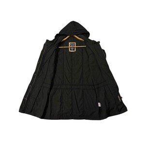 Puede incluir: Chaqueta negra con capucha y cremallera completa, abierta para mostrar el forro interior. La chaqueta tiene una cintura ajustable con cord&oacute;n y una peque&ntilde;a etiqueta cerca del cuello. Se muestra en una percha de madera.