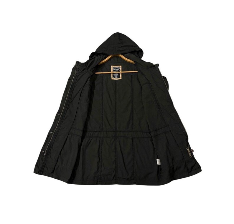 Puede incluir: Chaqueta negra con capucha y cremallera completa, abierta para mostrar el forro interior. La chaqueta tiene una cintura ajustable con cord&oacute;n y una peque&ntilde;a etiqueta cerca del cuello. Se muestra en una percha de madera.