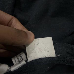 Puede incluir: Primer plano de una etiqueta de prenda azul marino oscuro con texto japon&eacute;s y s&iacute;mbolos de cuidado. La etiqueta est&aacute; sujeta por una mano. Tambi&eacute;n es visible una etiqueta blanca m&aacute;s peque&ntilde;a con la marca "chapeau" y "GARCE".