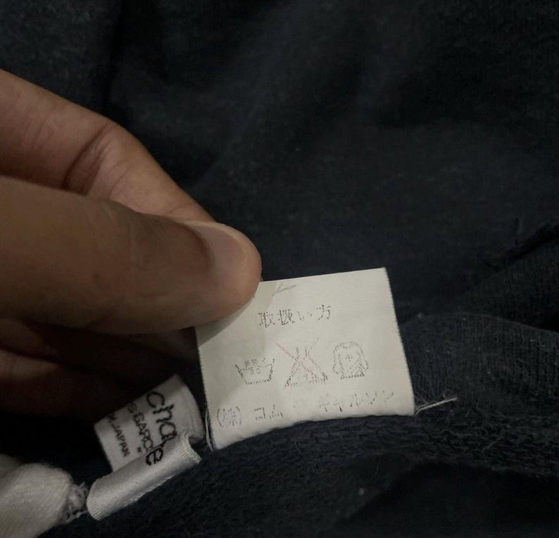 Puede incluir: Primer plano de una etiqueta de prenda azul marino oscuro con texto japon&eacute;s y s&iacute;mbolos de cuidado. La etiqueta est&aacute; sujeta por una mano. Tambi&eacute;n es visible una etiqueta blanca m&aacute;s peque&ntilde;a con la marca "chapeau" y "GARCE".