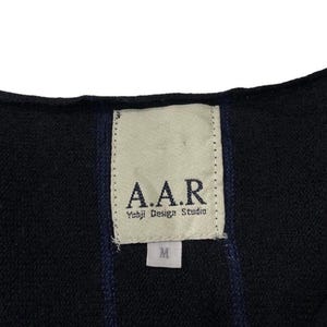 Puede incluir: Primer plano de una prenda negra con rayas verticales azul marino. Una etiqueta rectangular est&aacute; cosida, mostrando el texto "A.A.R. Yohji Design Studio" y una talla "M". La tela parece ser un material de punto o tejido.