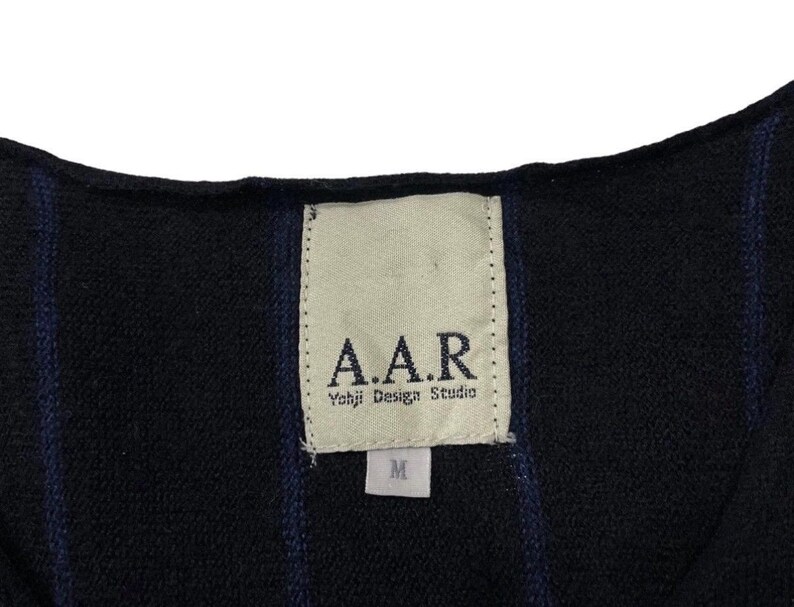 Puede incluir: Primer plano de una prenda negra con rayas verticales azul marino. Una etiqueta rectangular est&aacute; cosida, mostrando el texto "A.A.R. Yohji Design Studio" y una talla "M". La tela parece ser un material de punto o tejido.