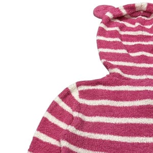 May include: A pink and white striped hooded sweater with a fuzzy texture. The hood features a small, rounded ear detail at the top. The sweater's stripes are horizontal and evenly spaced.
