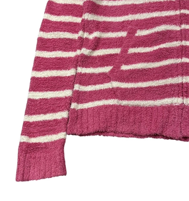May include: Close-up of a pink and white striped sweater. The sweater has a soft, textured appearance and a horizontal stripe pattern. The sleeve and body are visible, showing the knit fabric.