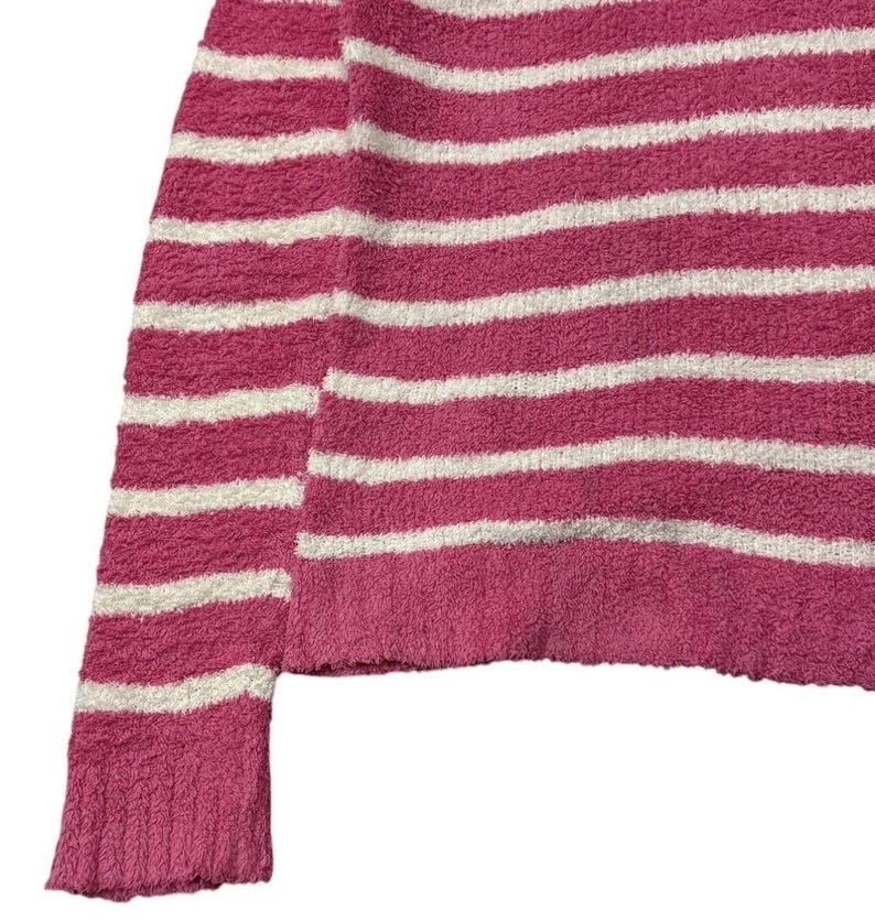 May include: Close-up of a pink and white striped sweater. The sweater has horizontal stripes and a textured, fuzzy appearance. The sleeve and hem are visible, showcasing the garment's construction and color scheme.
