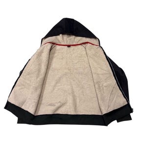 May include: Black hooded zip-up jacket with a soft, cream-coloured fleece lining. The hood is lined with the same fleece material. A red trim is visible along the inside collar. The jacket is open, displaying the interior.