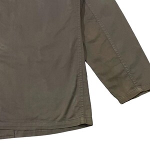 May include: Close-up of a khaki-colored jacket. The jacket has a button-up front and a three-quarter length sleeve. The fabric appears to be a durable cotton or blend, with visible stitching details along the seams.