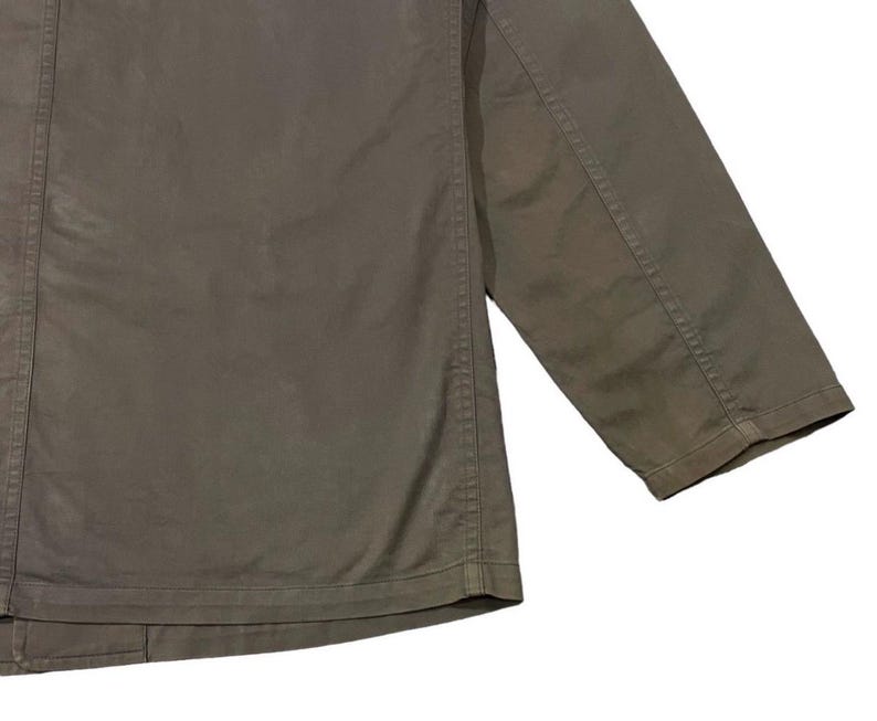 May include: Close-up of a khaki-colored jacket. The jacket has a button-up front and a three-quarter length sleeve. The fabric appears to be a durable cotton or blend, with visible stitching details along the seams.