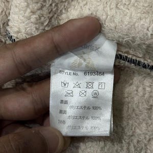 May include: A close-up of a clothing label with the text "STYLE No. 6193464". The label displays care symbols and fabric composition details. The fabric is 100% polyester. The background is a textured, beige fabric.