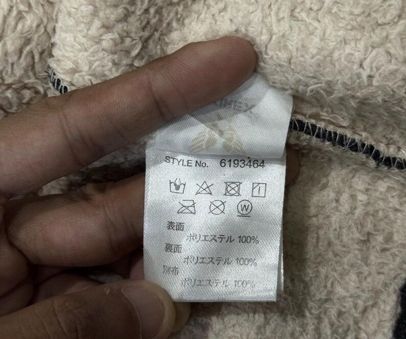 May include: A close-up of a clothing label with the text "STYLE No. 6193464". The label displays care symbols and fabric composition details. The fabric is 100% polyester. The background is a textured, beige fabric.