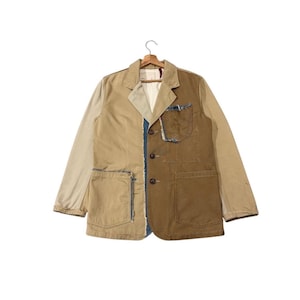 May include: A two-tone blazer in tan and brown, with a patchwork design. The jacket features a notched lapel, three buttons, and two front pockets. The seams are accented with blue denim. The blazer is hanging on a wooden hanger.