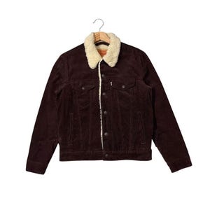 Op de afbeelding: Een donkerbruine corduroy jas met een crèmekleurige shearling kraag en voering. De jas heeft knoopsluitingen, twee borstzakken en twee zijzakken. De jas hangt aan een houten kleerhanger.