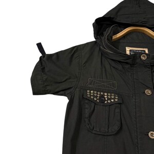 Puede incluir: Chaqueta verde oliva oscuro con capucha y mangas cortas. La chaqueta tiene un bolsillo con tachuelas met&aacute;licas y botones de madera. La chaqueta est&aacute; colgada en una percha de madera.