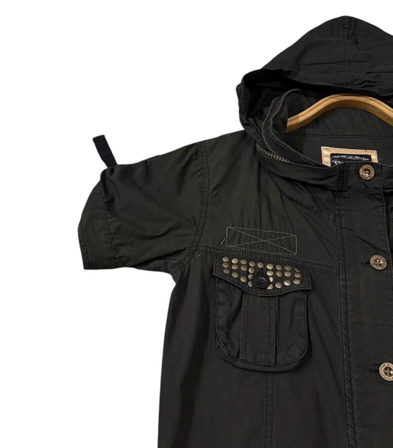 Puede incluir: Chaqueta verde oliva oscuro con capucha y mangas cortas. La chaqueta tiene un bolsillo con tachuelas met&aacute;licas y botones de madera. La chaqueta est&aacute; colgada en una percha de madera.