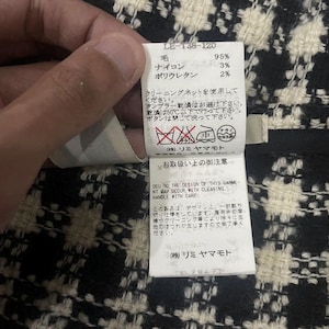 May include: A close-up of a clothing tag with Japanese text and care symbols. The tag is white with black text and is held by a hand. The fabric has a black and white checkered pattern.