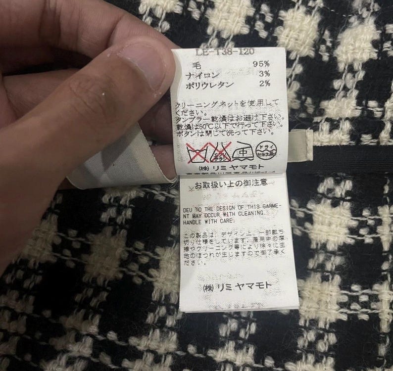 May include: A close-up of a clothing tag with Japanese text and care symbols. The tag is white with black text and is held by a hand. The fabric has a black and white checkered pattern.