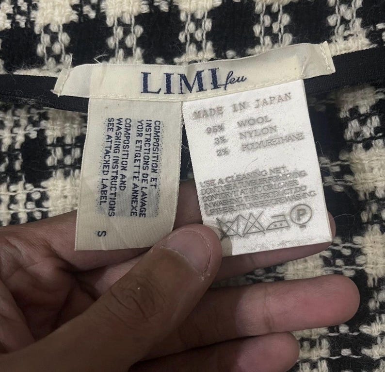 May include: Close-up of a black and white woven fabric with a tag that reads "LIMI feu" and "MADE IN JAPAN." The tag lists the fabric composition as 95% wool, 3% nylon, and 2% polyurethane. Washing instructions are also included.