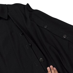 May include: A black coat with a collar and button closures. The coat is hanging on a wooden hanger. The coat has buttons on the sleeves and down the front.