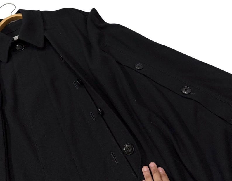 May include: A black coat with a collar and button closures. The coat is hanging on a wooden hanger. The coat has buttons on the sleeves and down the front.